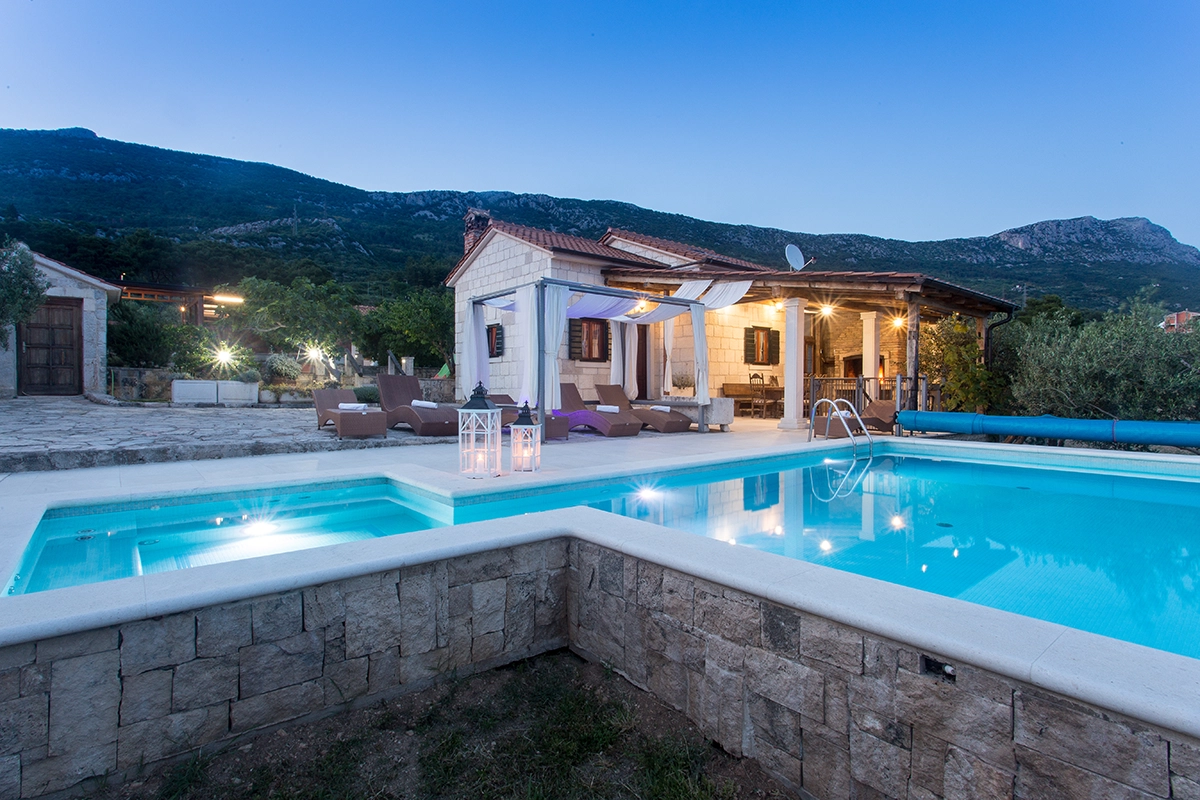 View of the illuminated pool, villa, and mountain at dusk, featuring the front side of the villa, the pool with lounge chairs, and the house's entrance area.